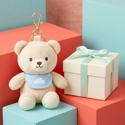 Cream plush bear charm with blue cloud bib beside ivory box with mint ribbon