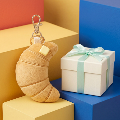 Golden crescent croissant plush charm with butter accent beside unbranded ivory mint-ribbon box